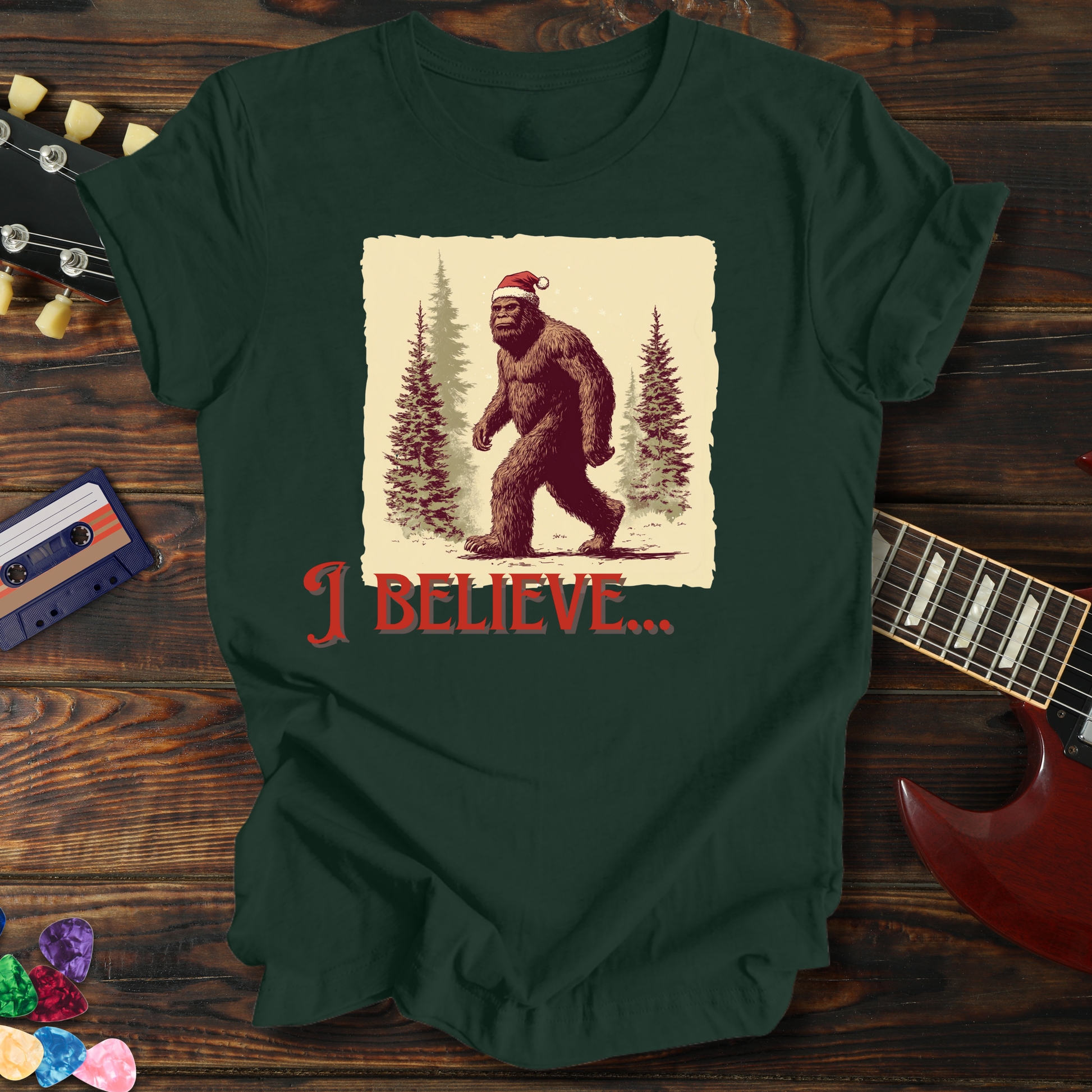 Green t-shirt with a Christmas-themed Bigfoot graphic and 'I Believe' text on a wooden surface with a guitar and cassette tape.
