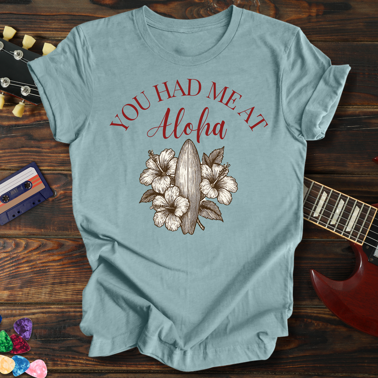 Light blue t-shirt with 'You Had Me At Aloha' text and floral design on a wooden surface with a guitar and cassette tape.