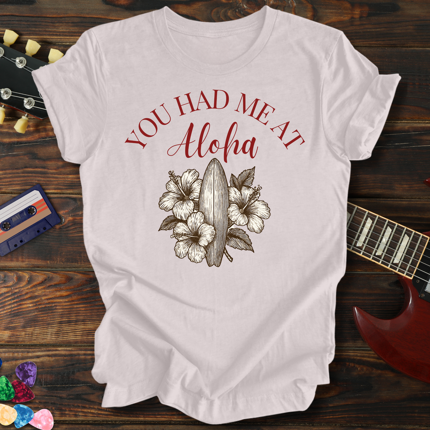 White t-shirt with 'You Had Me At Aloha' design on a wooden surface with a guitar and cassette tape.