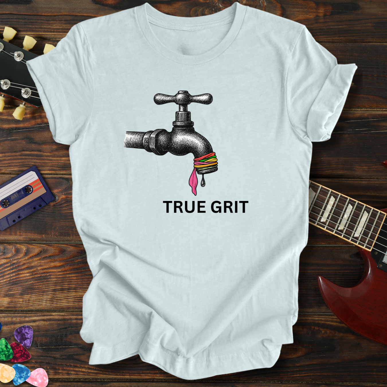 White t-shirt with a graphic of a faucet and colorful beads, text 'TRUE GRIT', on a wooden surface with a guitar and cassette tape.