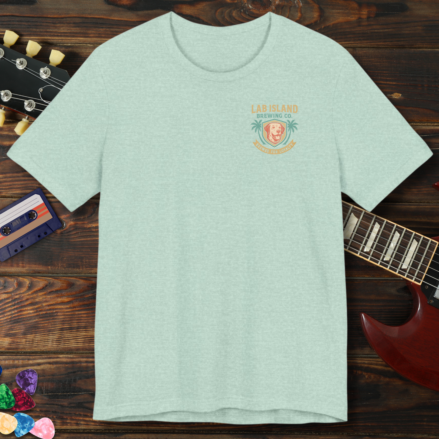 Light green t-shirt with a logo on a wooden surface with a guitar and cassette tape.