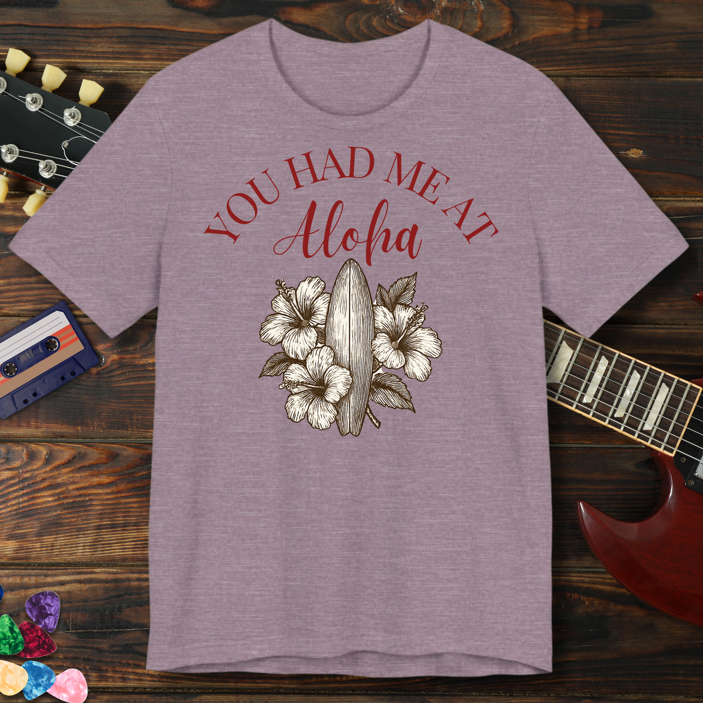 Purple t-shirt with 'You Had Me At Aloha' text and floral design on a wooden background with a guitar and cassette tape.
