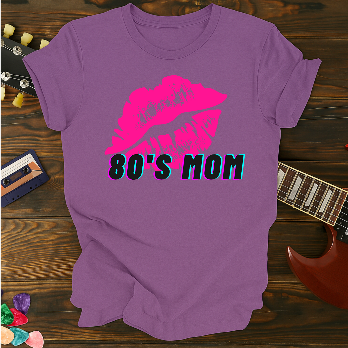 Purple t-shirt with pink lips and '80s Mom' text on a wooden surface with a guitar and cassette tape.