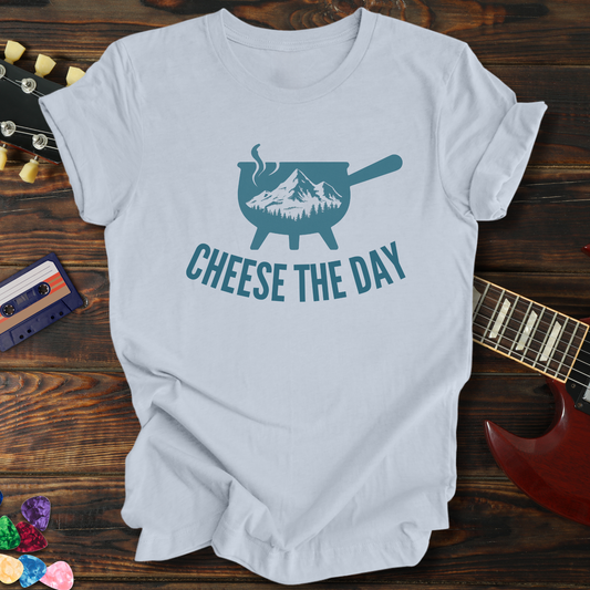 White t-shirt with 'Cheese the Day' graphic design on a wooden surface with a guitar and colorful objects.