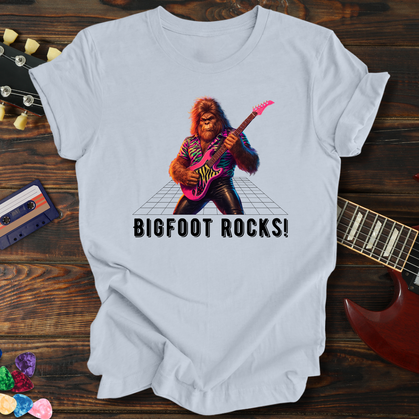 White t-shirt with a graphic of a rock-playing bigfoot and 'BIGFOOT ROCKS!' text, on a wooden surface with guitar and cassette tape.