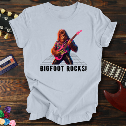 White t-shirt with a graphic of a rock-playing bigfoot and 'BIGFOOT ROCKS!' text, on a wooden surface with guitar and cassette tape.