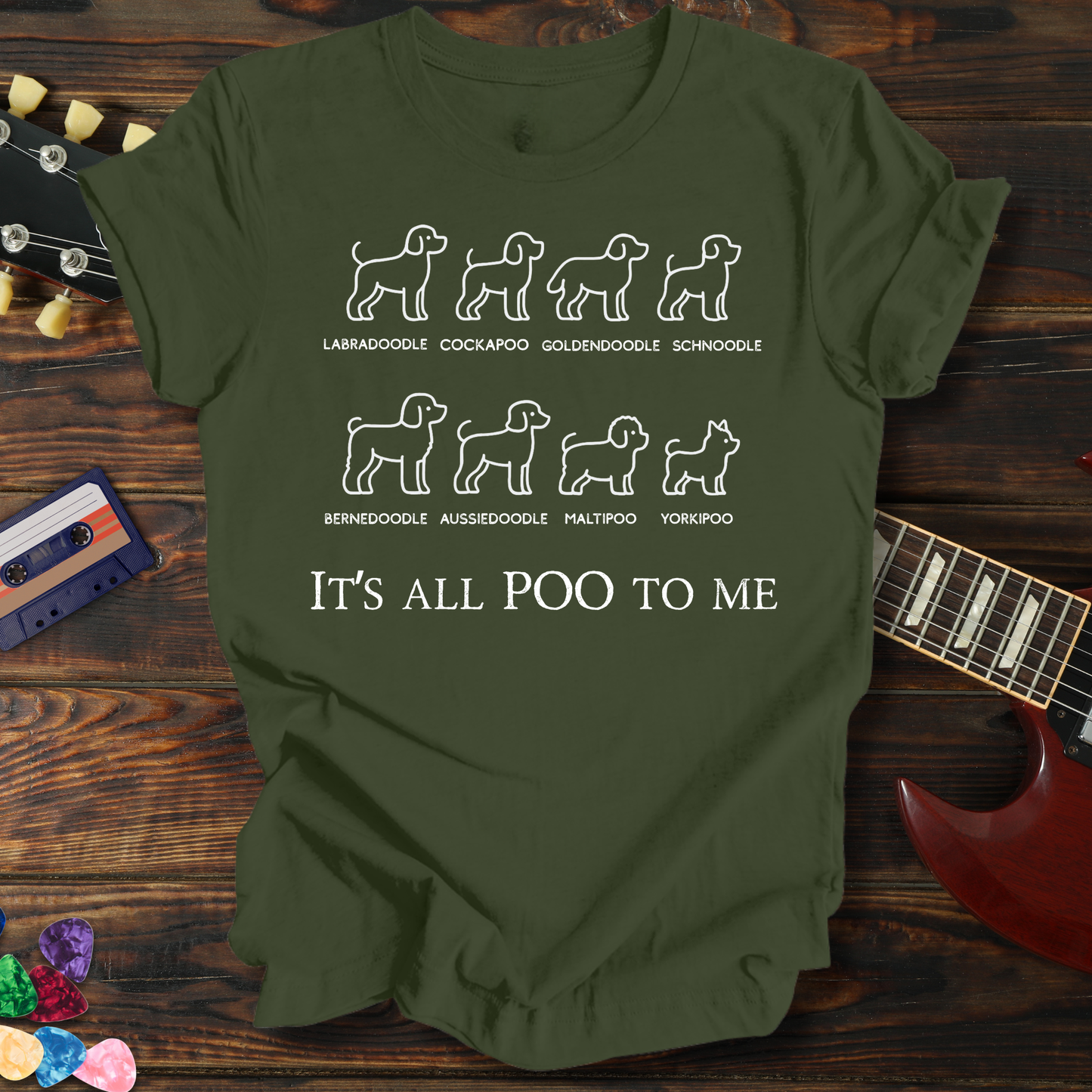 Green t-shirt with dog breed illustrations and text on a wooden surface with a guitar and cassette tape.
