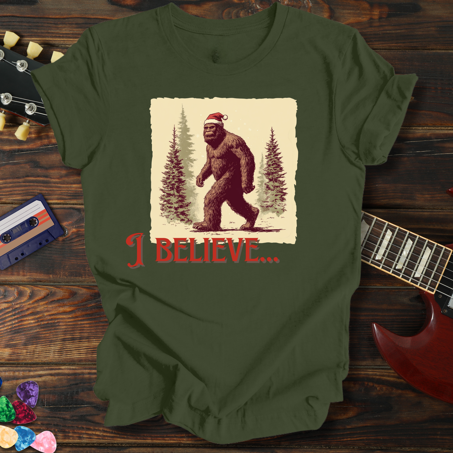 Green t-shirt with a graphic of a bear and 'I Believe' text on a wooden surface with a guitar and cassette tape.
