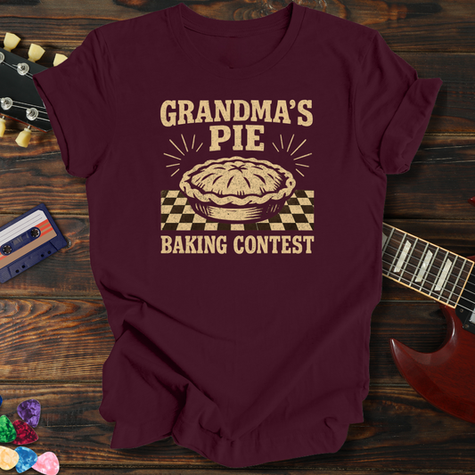 Maroon t-shirt with 'Grandma's Pie Baking Contest' design on a wooden surface with a guitar and cassette tape.