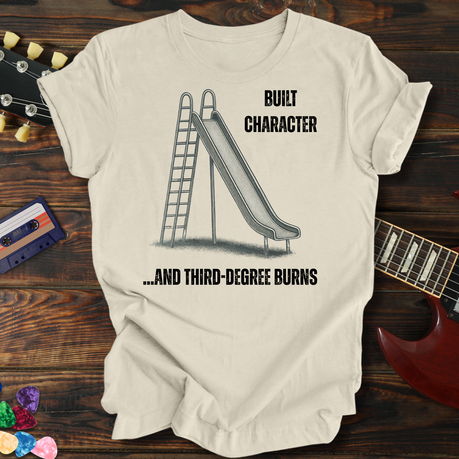 Beige t-shirt with graphic design and text on a wooden surface with a guitar and cassette tape.
