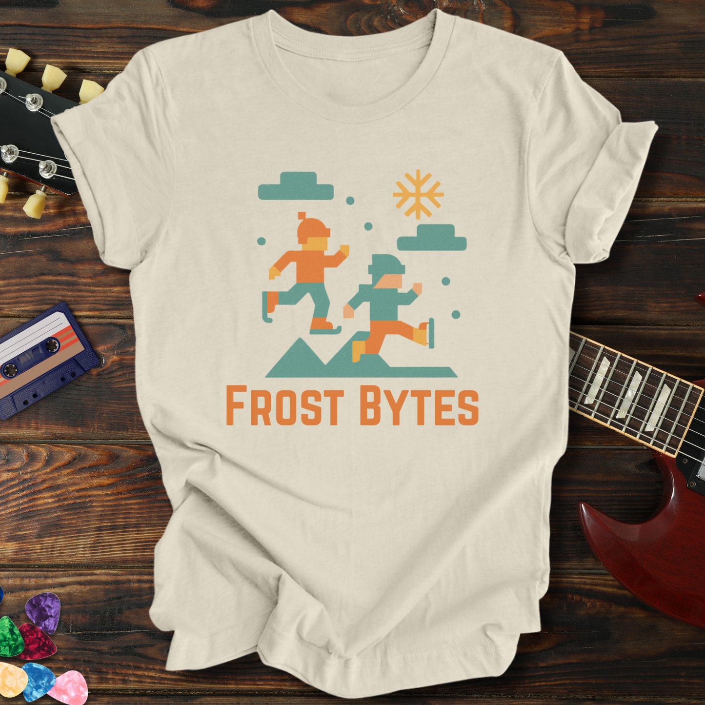 Beige t-shirt with 'Frost Bytes' graphic design on a wooden surface with a guitar and cassette tape.