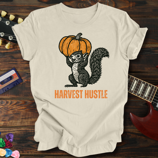 Beige t-shirt with a graphic of a squirrel with a pumpkin and 'Harvest Hustle' text, placed on a wooden surface with a guitar and cassette tape.