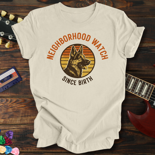 White t-shirt with 'Neighborhood Watch Since Birth' graphic on a wooden surface with a guitar and cassette tape.