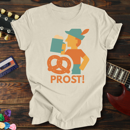 Beige t-shirt with a graphic design of a person holding a pretzel and the word 'PROST!' on a wooden surface with a guitar and cassette tape.