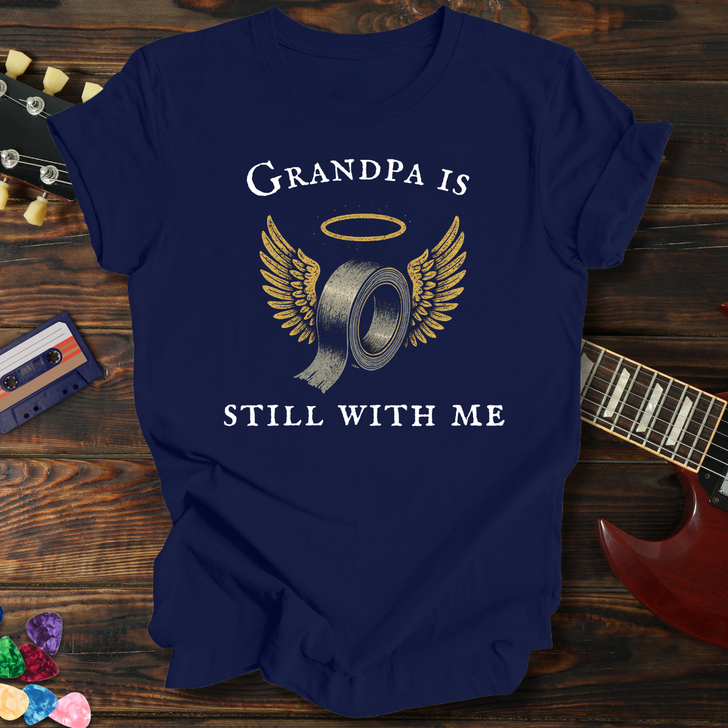 Navy blue t-shirt with 'Grandpa is still with me' graphic on a wooden surface with a guitar and cassette tape.