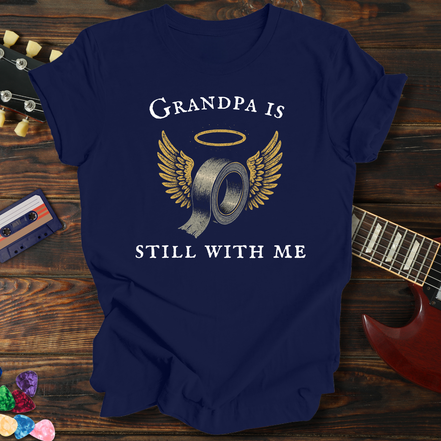 Navy blue t-shirt with 'Grandpa is still with me' graphic on a wooden surface with a guitar and cassette tape.