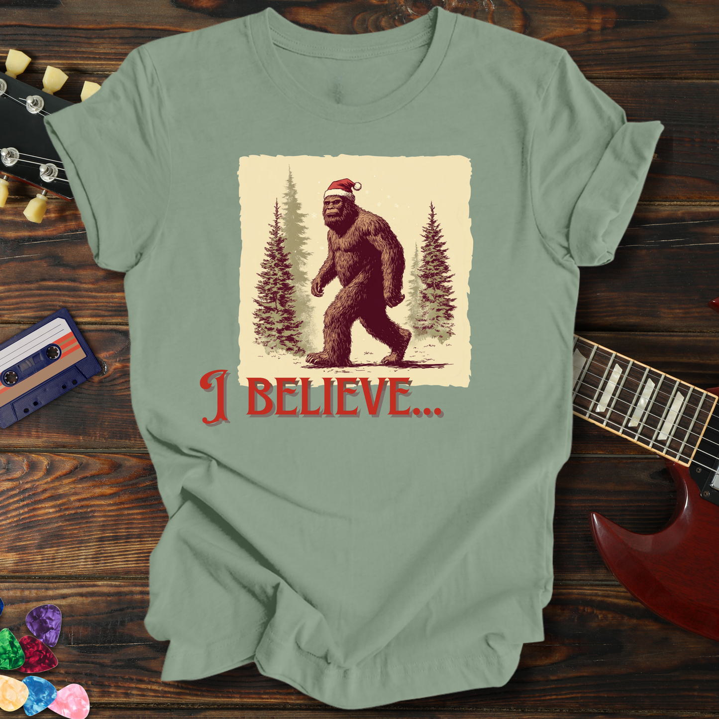 Green t-shirt with a graphic of a bear and 'I Believe' text on a wooden surface with a guitar and cassette tape.