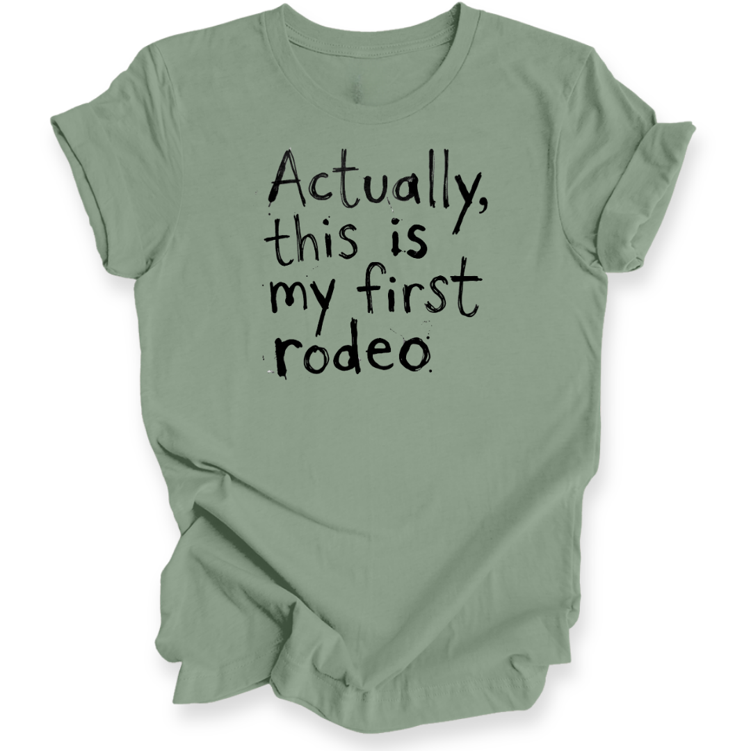 A sage green short-sleeve t-shirt with the phrase 'Actually, this is my first rodeo' printed in a black, hand-written style font on the front, displayed against a clean white background.