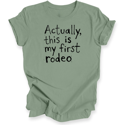 A sage green short-sleeve t-shirt with the phrase 'Actually, this is my first rodeo' printed in a black, hand-written style font on the front, displayed against a clean white background.