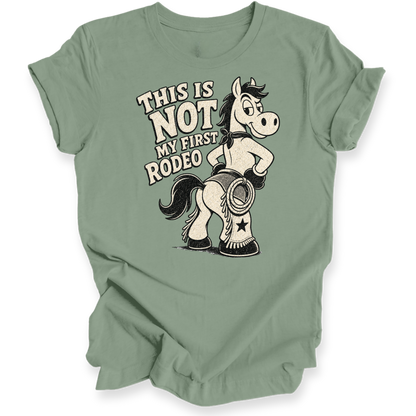 A sage green short-sleeve t-shirt with a graphic of a cartoon horse wearing cowboy boots and the text 'THIS IS NOT MY FIRST RODEO' printed on the front, displayed against a plain white background.