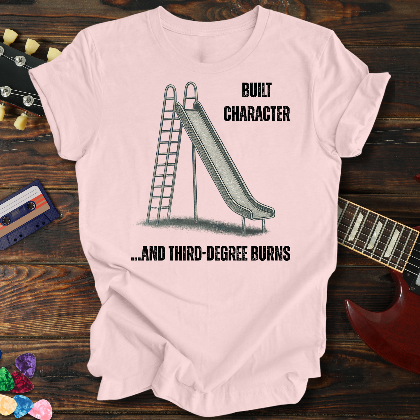 Pink t-shirt with a graphic of a slide and text on a wooden surface with a guitar and cassette tape.