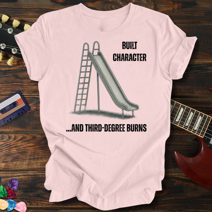 Pink t-shirt with a graphic of a slide and text on a wooden surface with a guitar and cassette tape.