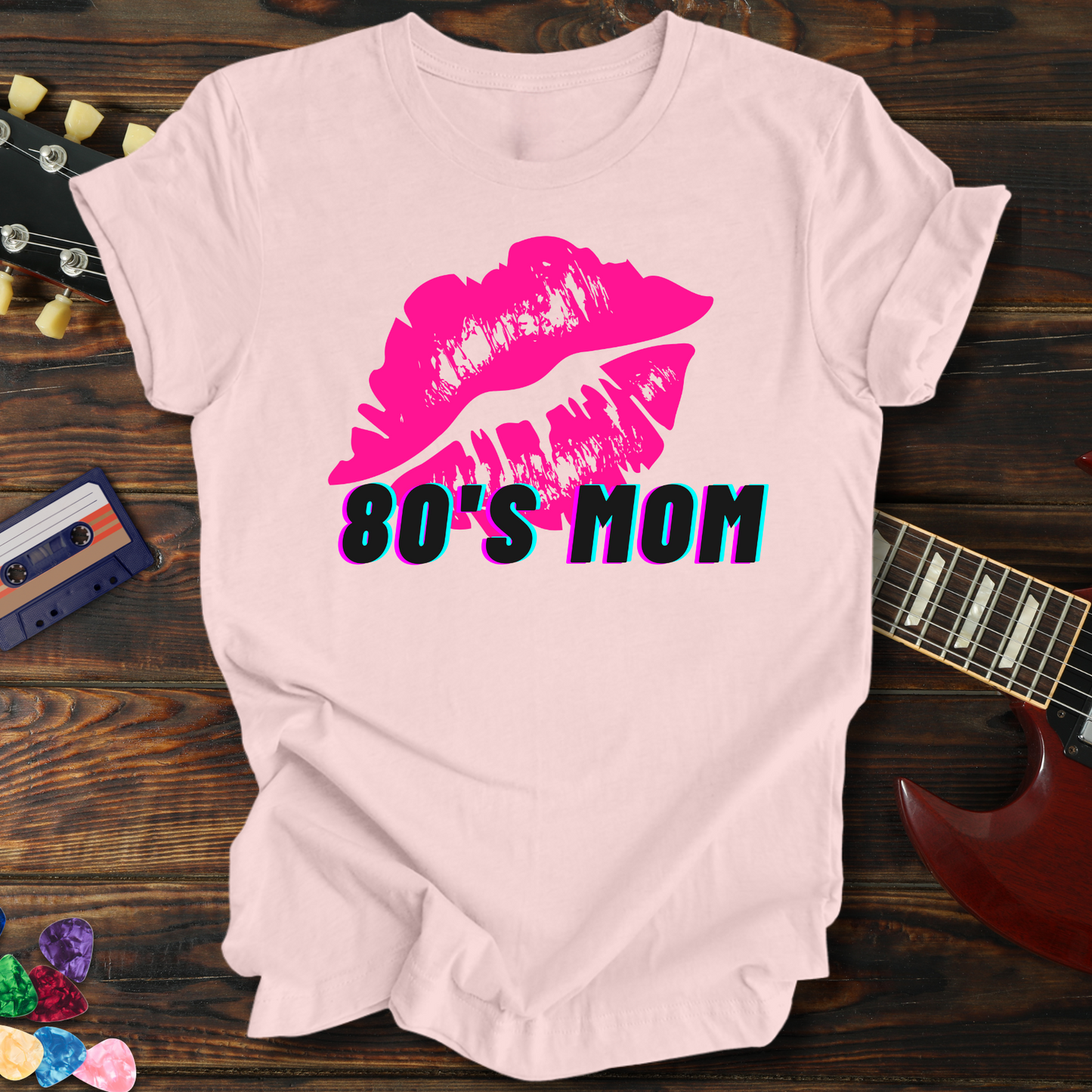 Pink t-shirt with pink lips and '80's Mom' text on a wooden surface with a guitar and cassette tape.