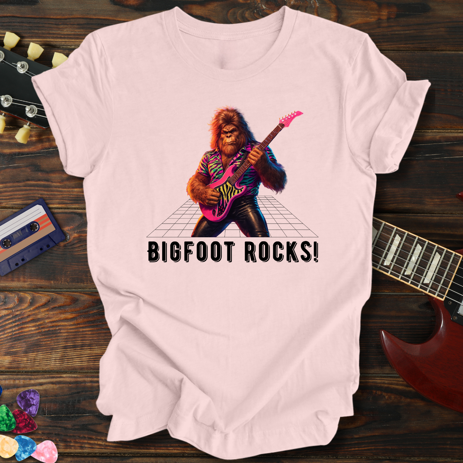 Pink t-shirt with a graphic of a bigfoot playing guitar and 'BIGFOOT ROCKS!' text, on a wooden surface with a guitar and cassette tape.