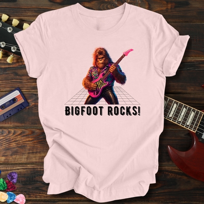 Pink t-shirt with a graphic of a bigfoot playing guitar and 'BIGFOOT ROCKS!' text, on a wooden surface with a guitar and cassette tape.