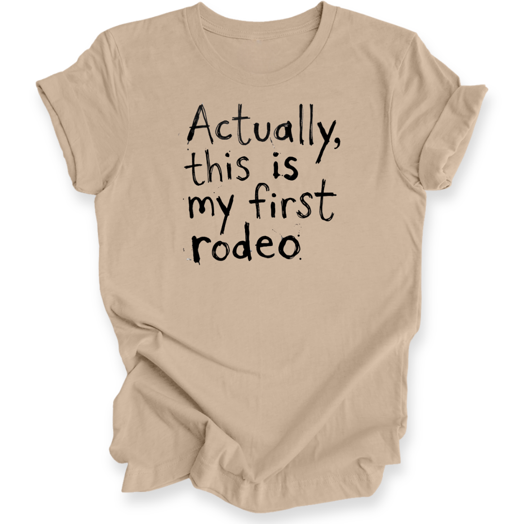 A beige short-sleeve t-shirt with black hand-lettered text reading 'Actually, this is my first rodeo' on the front, shown against a plain white background.