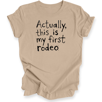A beige short-sleeve t-shirt with black hand-lettered text reading 'Actually, this is my first rodeo' on the front, shown against a plain white background.