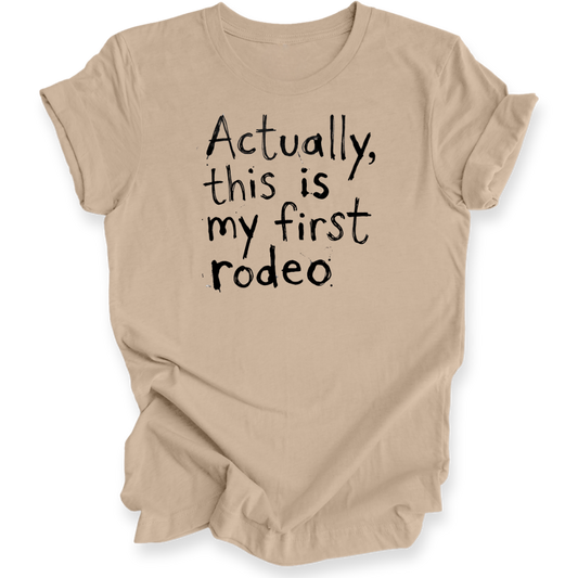 A beige short-sleeve t-shirt with black hand-lettered text reading 'Actually, this is my first rodeo' on the front, shown against a plain white background.