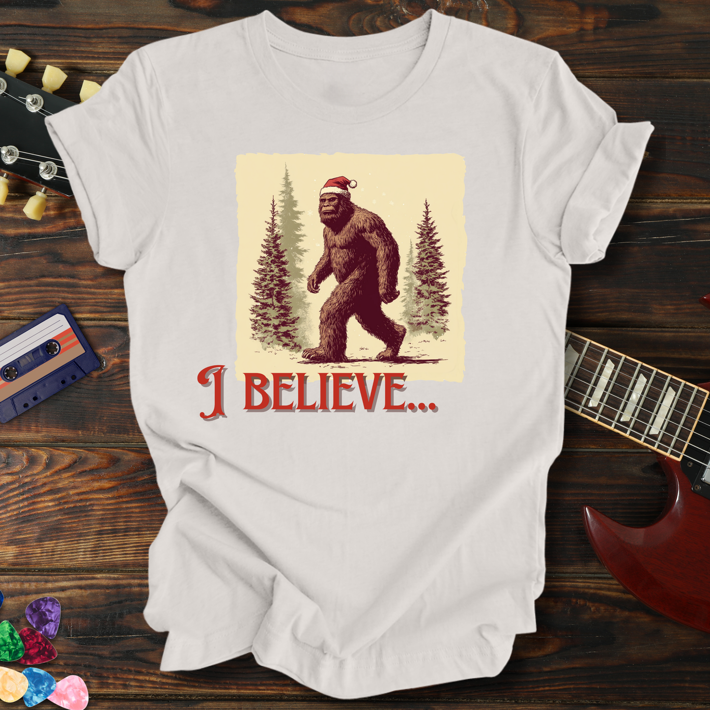 White t-shirt with a Christmas-themed Bigfoot graphic on a wooden surface with a guitar and cassette tape.