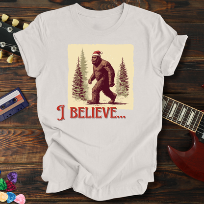 White t-shirt with a Christmas-themed Bigfoot graphic on a wooden surface with a guitar and cassette tape.