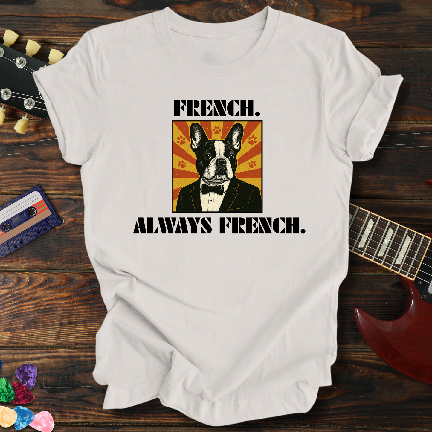 White t-shirt with a dog graphic and text 'FRENCH. ALWAYS FRENCH.' on a wooden surface with a guitar and cassette tape.