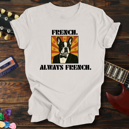 White t-shirt with a dog graphic and text 'FRENCH. ALWAYS FRENCH.' on a wooden surface with a guitar and cassette tape.