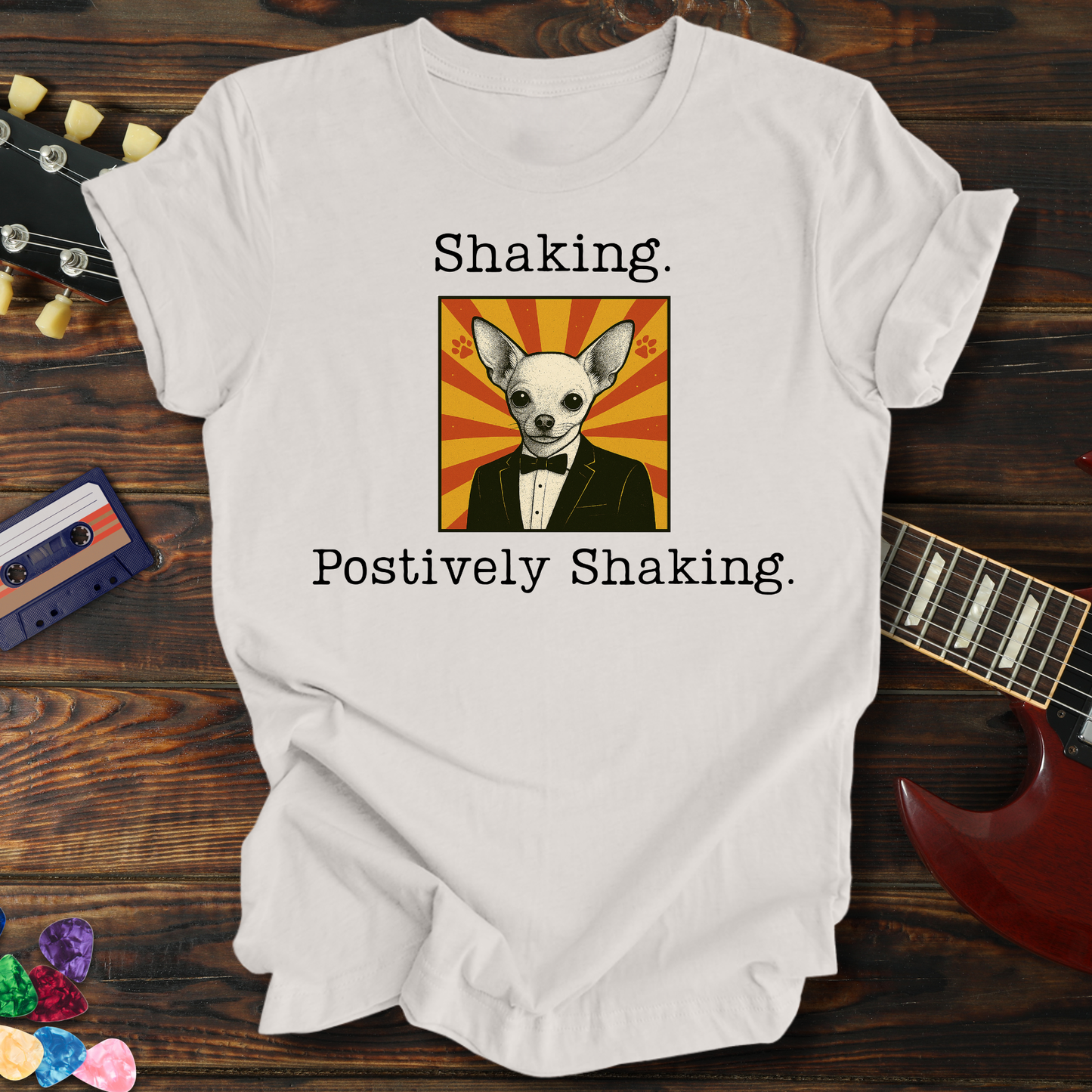 White t-shirt with a dog graphic and text on a wooden surface with a guitar and cassette tape.