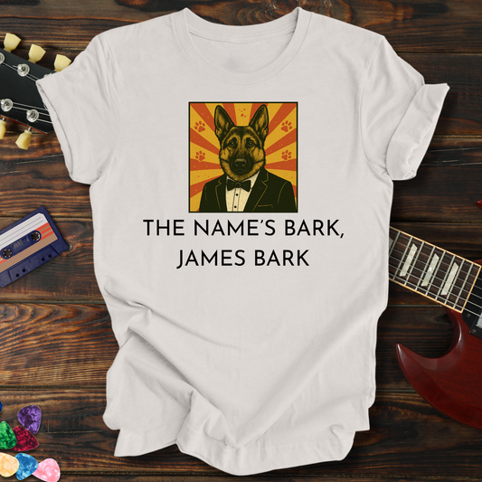 White t-shirt with a dog in a suit graphic and text 'The Name's Bark, James Bark' on a wooden surface with a guitar and cassette tape.