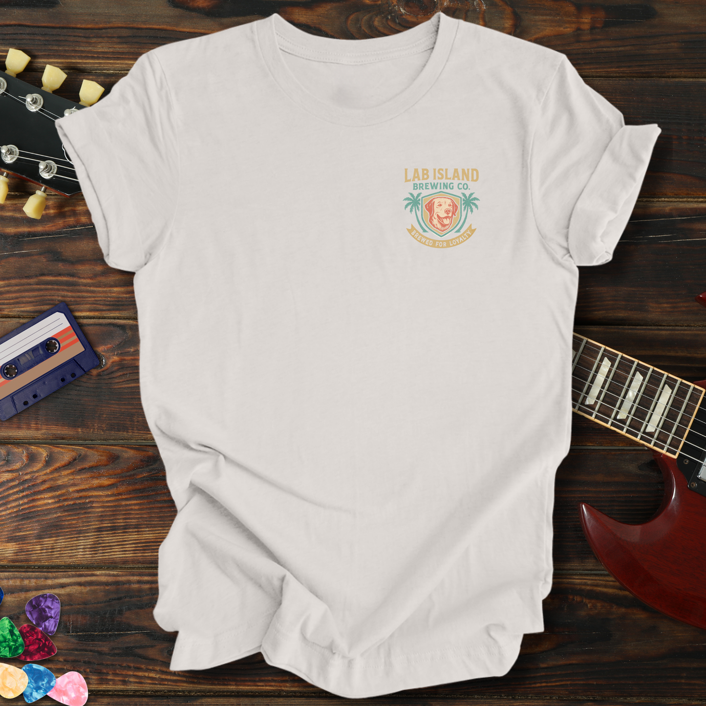 White t-shirt with a logo on a wooden surface with a guitar and cassette tape.