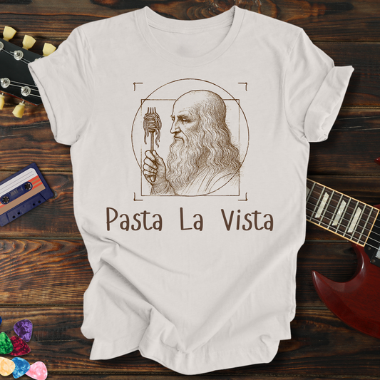 White t-shirt with Leonardo da Vinci holding a fork and 'Pasta La Vista' text on a wooden background.