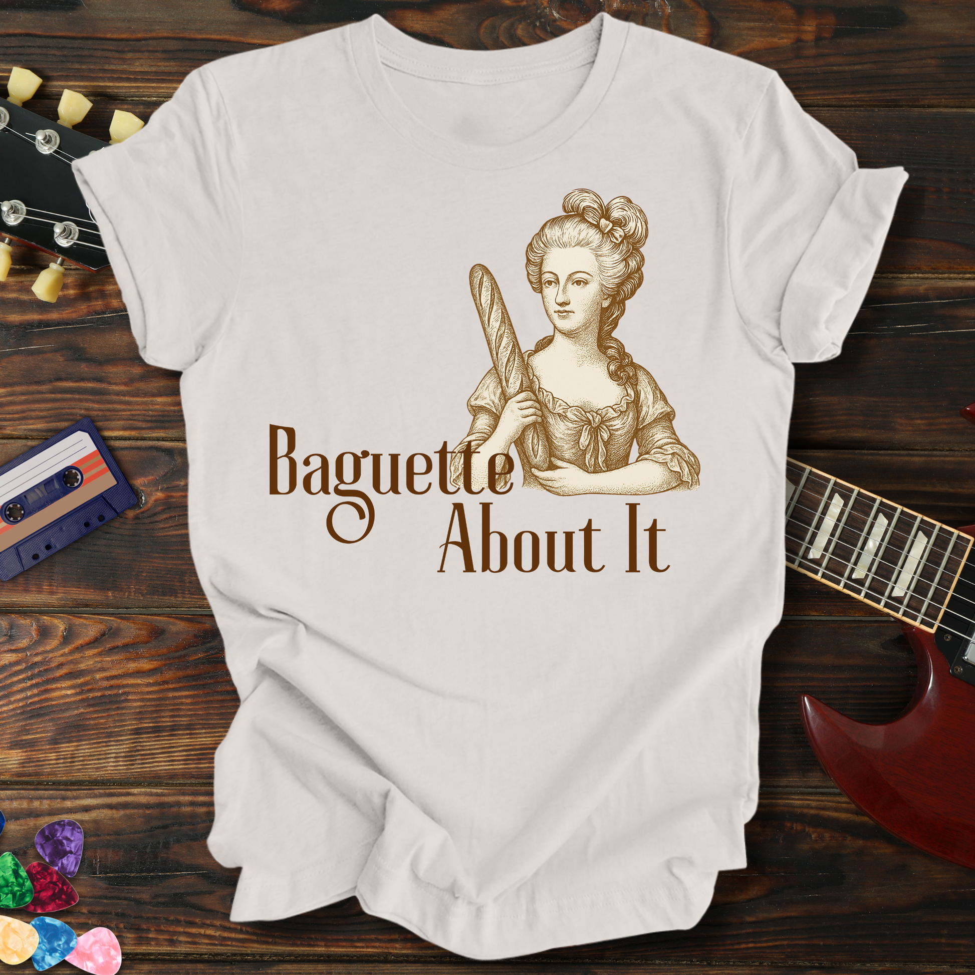 White t-shirt with a graphic of a woman holding a baguette and text 'Baguette About It' on a wooden background.