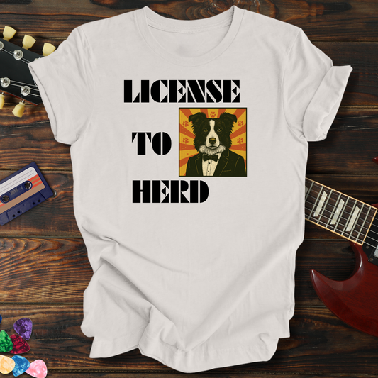 White t-shirt with a dog graphic and text 'LICENSE TO HERD' on a wooden surface with a guitar and cassette tape.