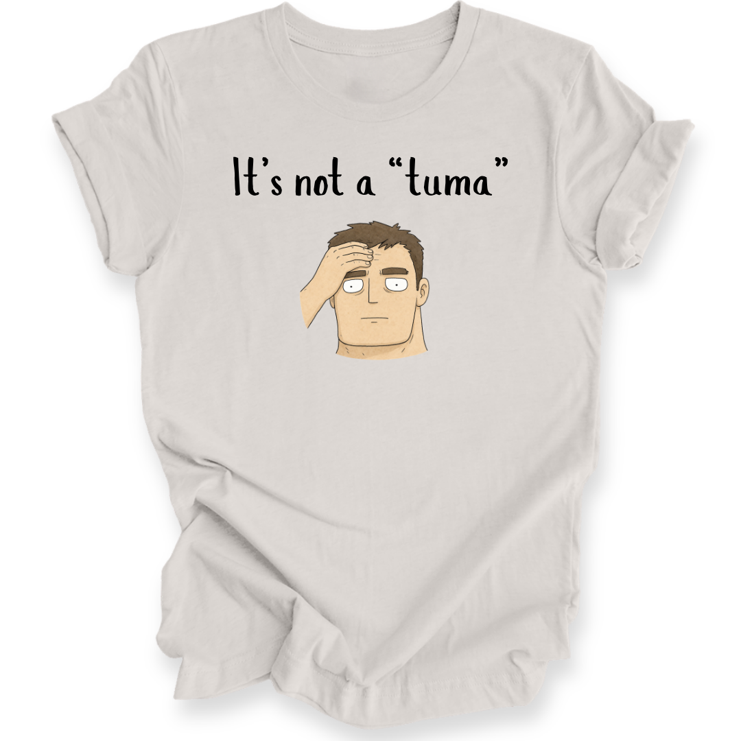 A light beige graphic t-shirt featuring the text 'It's not a "tuma"' above a cartoon illustration of a confused man with his hand on his head, displayed on a clean white background.
