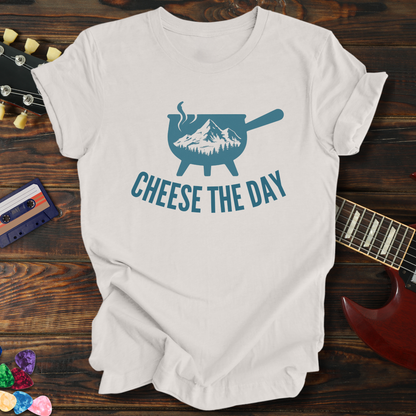 White t-shirt with 'Cheese the Day' graphic design on a wooden surface with a guitar and cassette tape.