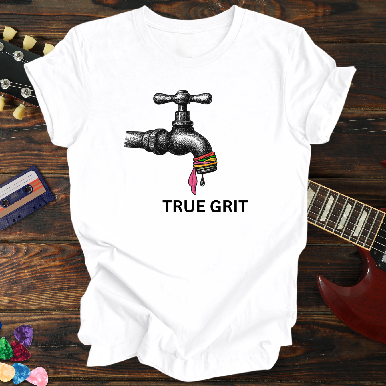 White t-shirt with a graphic of a faucet and colorful beads, text 'TRUE GRIT', on a wooden background with a guitar and cassette tape.