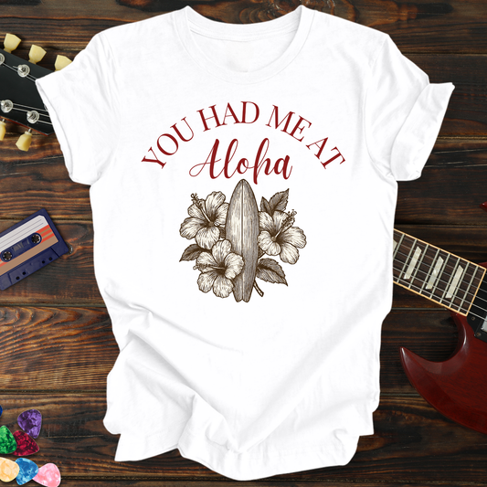 White t-shirt with 'You Had Me At Aloha' design on a wooden surface with a guitar and cassette tape.