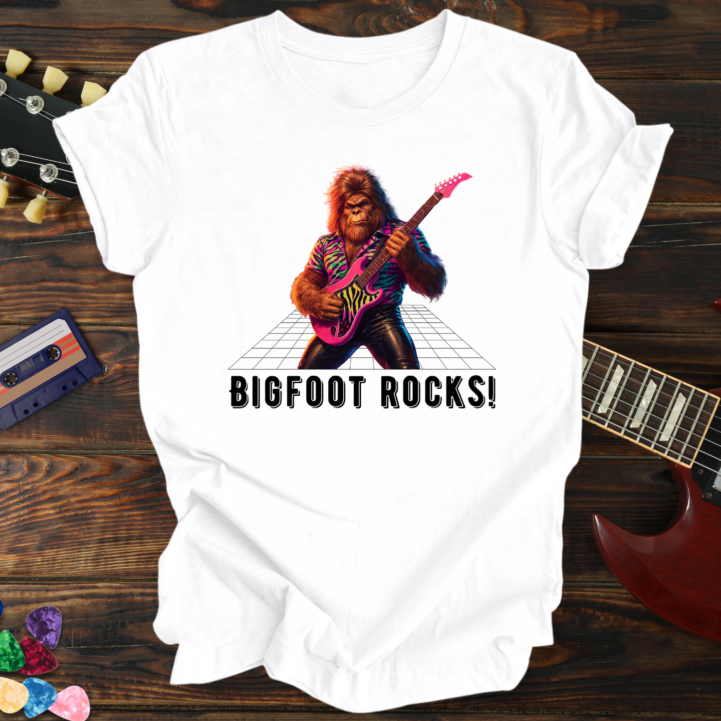 White t-shirt with a graphic of a bigfoot playing guitar and 'BIGFOOT ROCKS!' text, on a wooden surface with musical instruments.