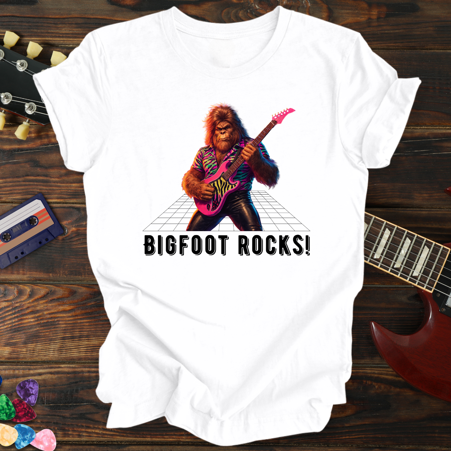 White t-shirt with a graphic of a bigfoot playing guitar and 'BIGFOOT ROCKS!' text, on a wooden surface with musical instruments.