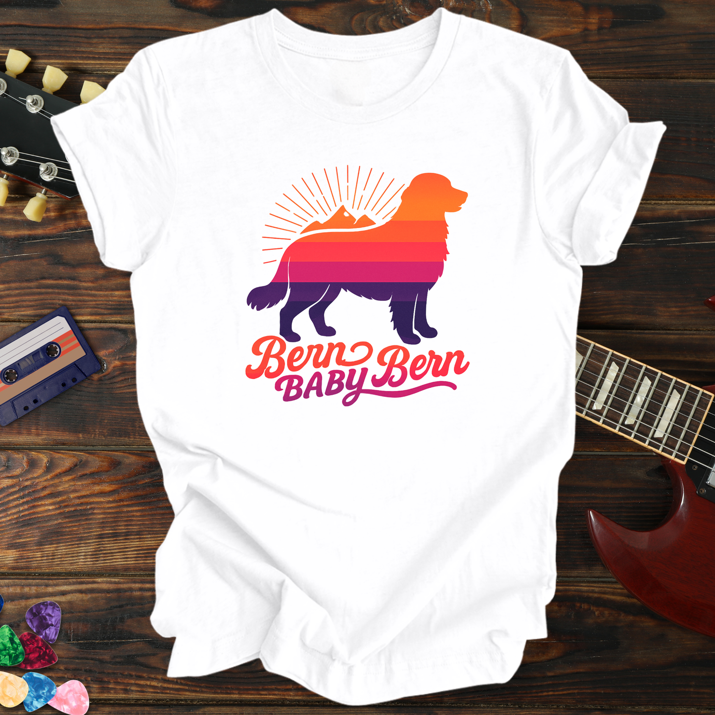 White t-shirt with colorful Bernese Mountain Dog design and 'Bern Baby Bern' text on a wooden surface with a guitar and cassette tape.