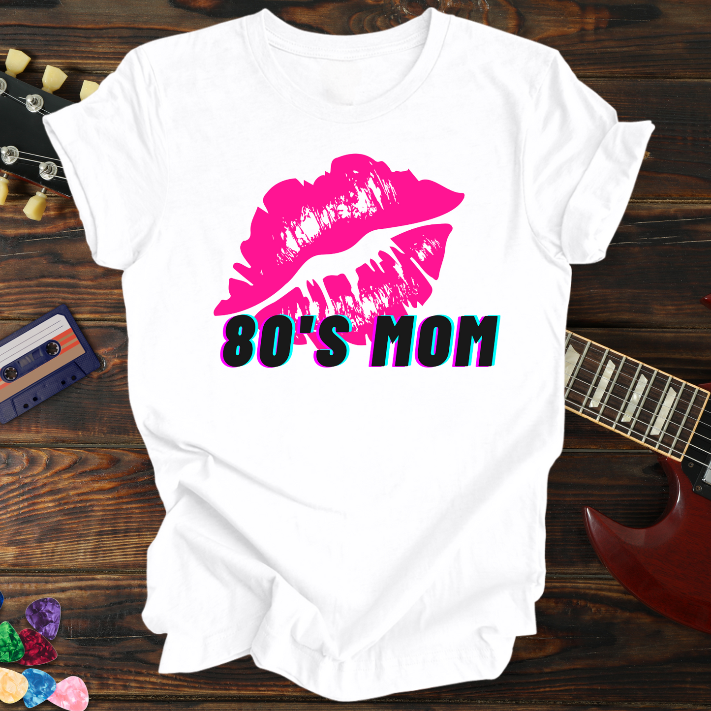 White t-shirt with pink lips and '80's Mom' text on a wooden surface with a guitar and cassette tape.
